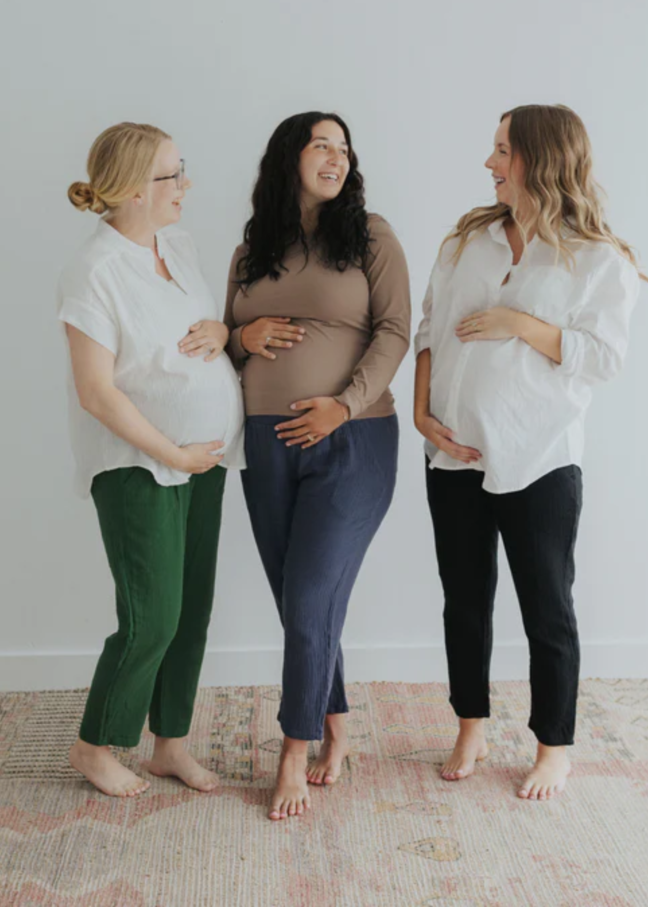 Three pregnant women smiling, wearing Everyday Breeze Pants in green, navy, and black cotton gauze.