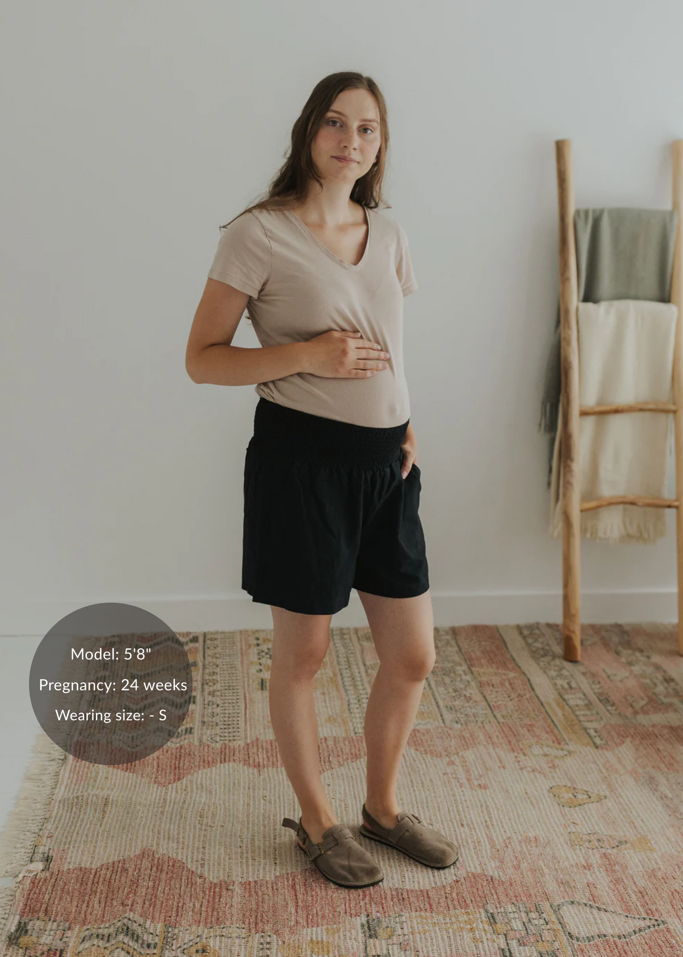 Pregnant woman in black linen smocked shorts and a beige tee, standing on a patterned rug.