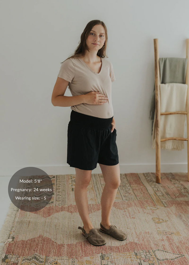 Pregnant woman in black linen smocked shorts and a beige tee, standing on a patterned rug.