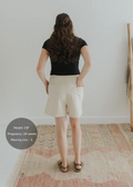 Woman from behind wearing natural linen smocked shorts with pockets and a black top.