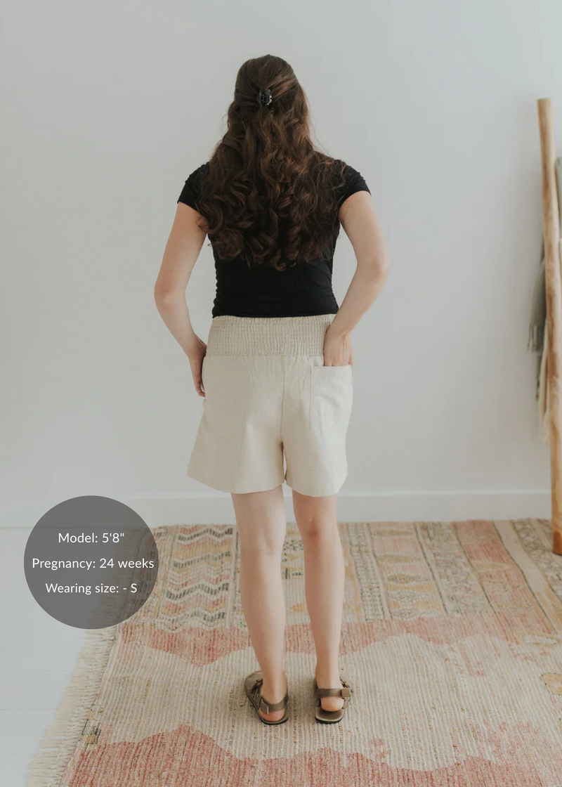 Woman from behind wearing natural linen smocked shorts with pockets and a black top.