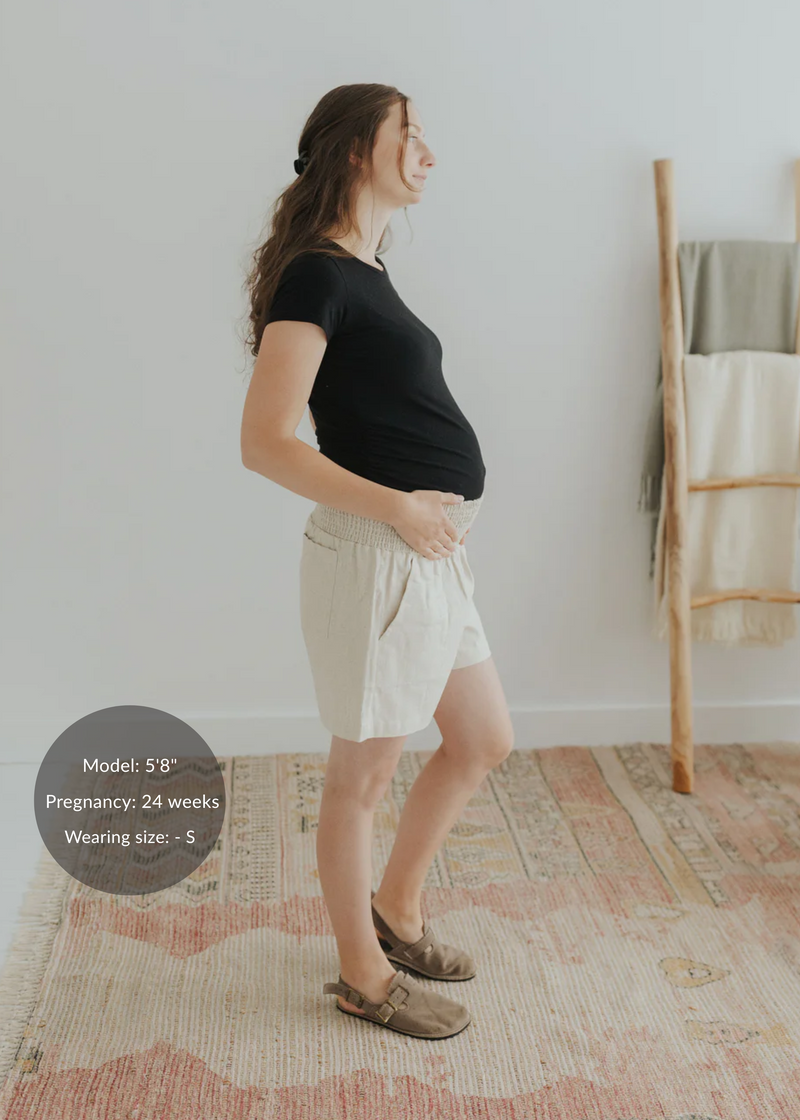 Pregnant woman in natural linen smocked shorts, black tee, and brown sandals.