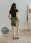 Woman from behind wearing olive green smocked linen shorts with deep pockets and a black top.