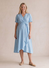 Smiling woman in a light blue adjustable wrap front dress with short sleeves and a tie waist.