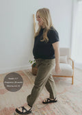 Smiling pregnant woman in olive cotton/linen side drawstring maternity pants and black tee, walking indoors.