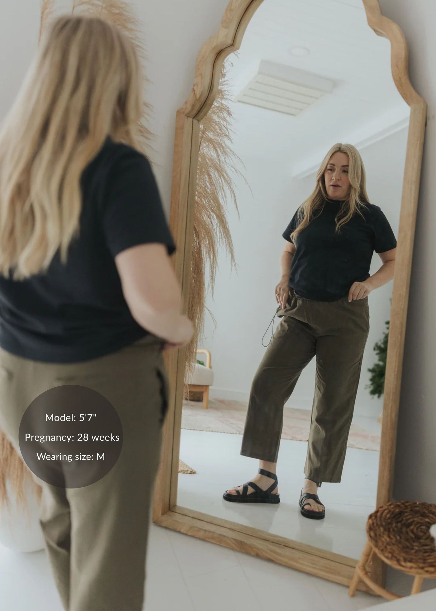 Blonde woman in olive cotton/linen side drawstring maternity pants and black top, reflected in a mirror.