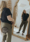 Smiling pregnant woman in a mirror wearing olive green cotton/linen side drawstring pants and a black top.