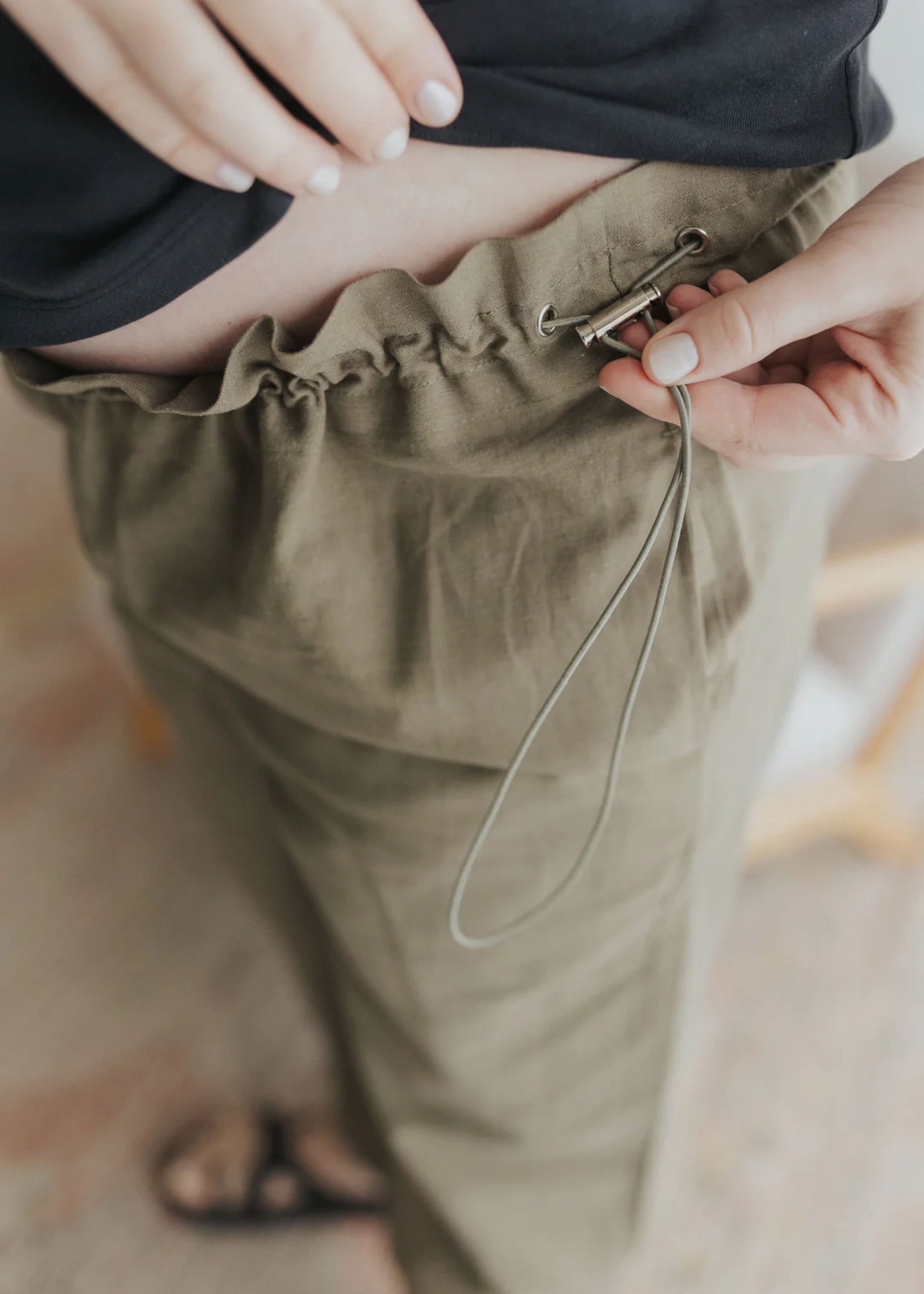 Close-up of a hand adjusting the side drawstring on olive cotton/linen maternity pants.