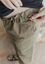 Close-up of a hand adjusting the side drawstring on olive cotton/linen maternity pants.