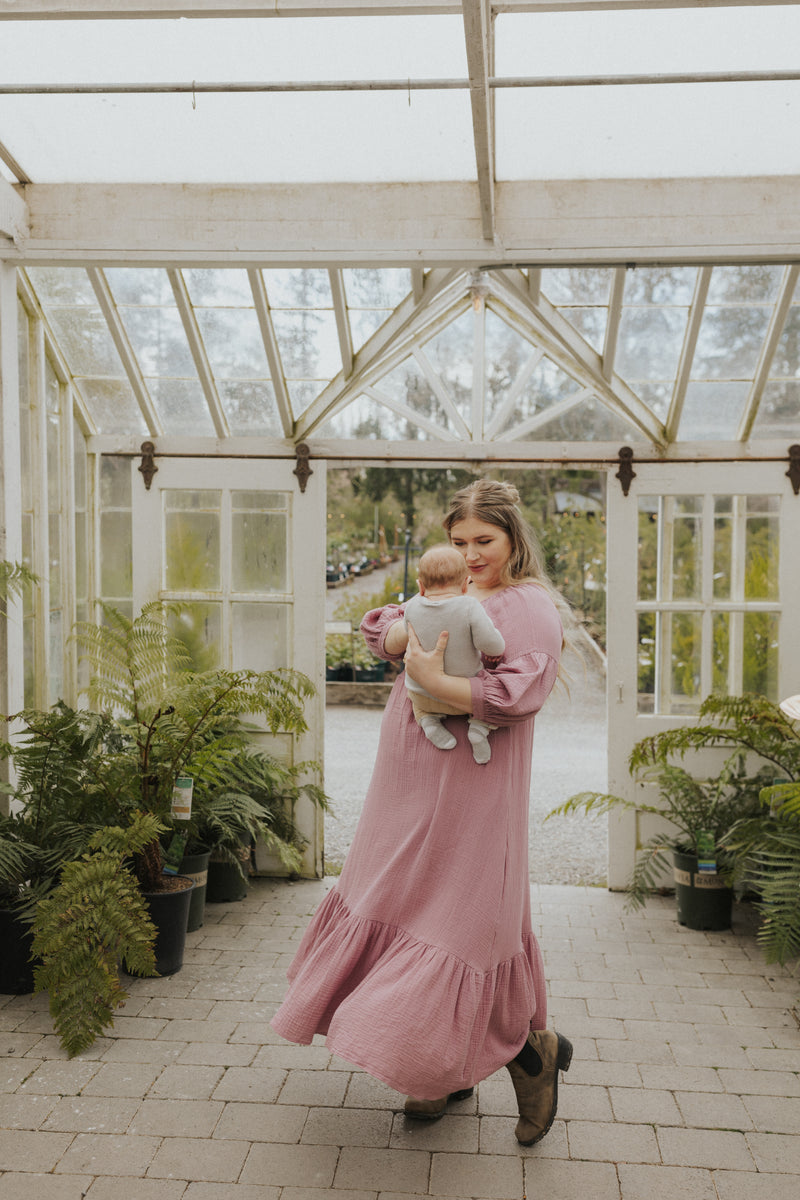 Woman in a pink cotton gauze Eliza maternity and nursing maxi dress with balloon sleeves, holding a baby.