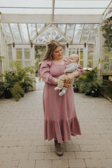 Woman in a pink Eliza Maternity and Nursing Dress in Cotton Gauze with balloon sleeves and ruffle skirt, holding a baby.
