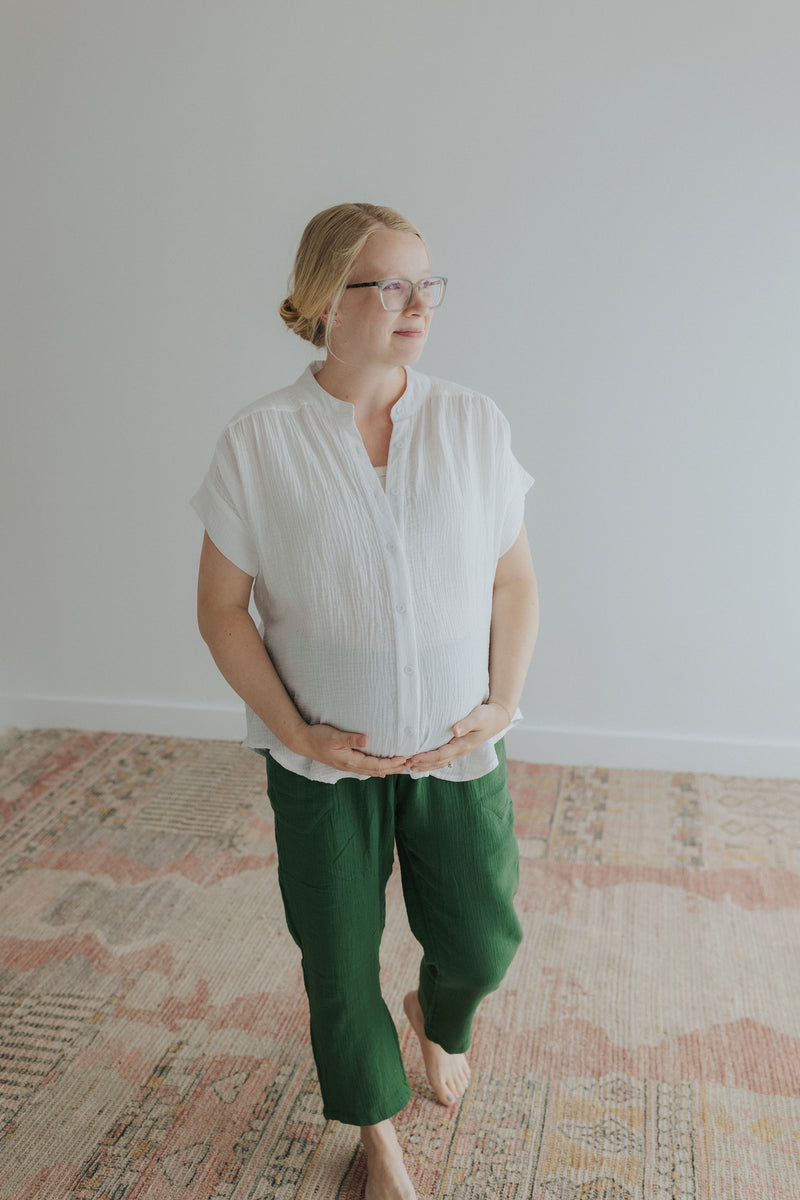Pregnant woman in a white cotton gauze button-down top and green pants, hands on her bump.