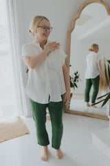 Smiling woman in white oversized cotton gauze button-down top and green pants, reflected in mirror.