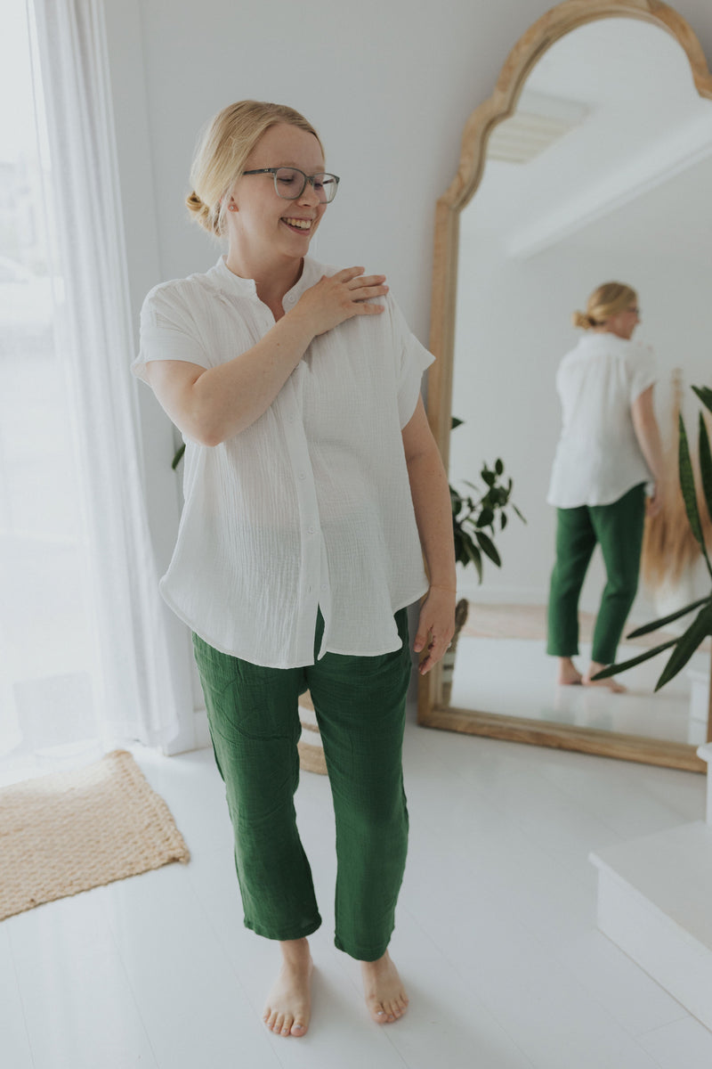 Smiling woman in white oversized cotton gauze button-down top and green pants, reflected in mirror.