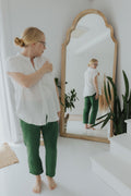 Woman in white cotton gauze button-down top and green pants, looking at her reflection in a wooden mirror.