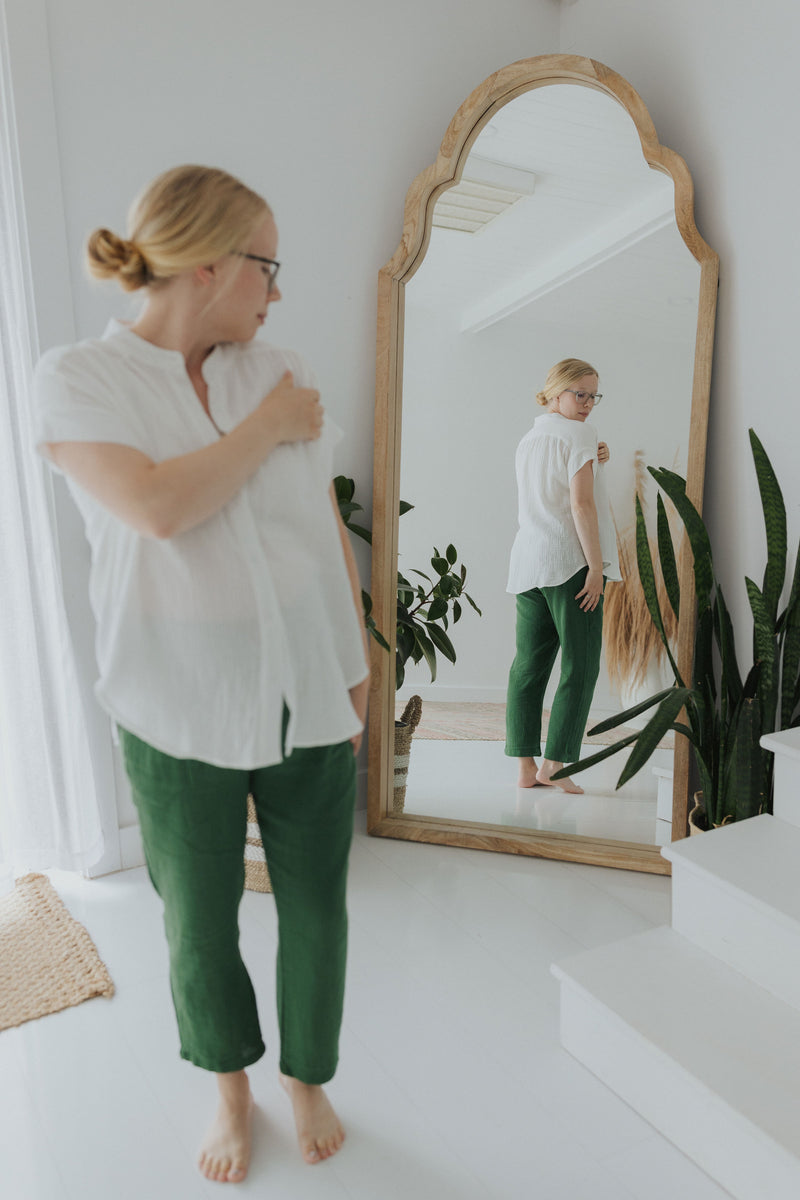 Woman in white cotton gauze button-down top and green pants, looking at her reflection in a wooden mirror.