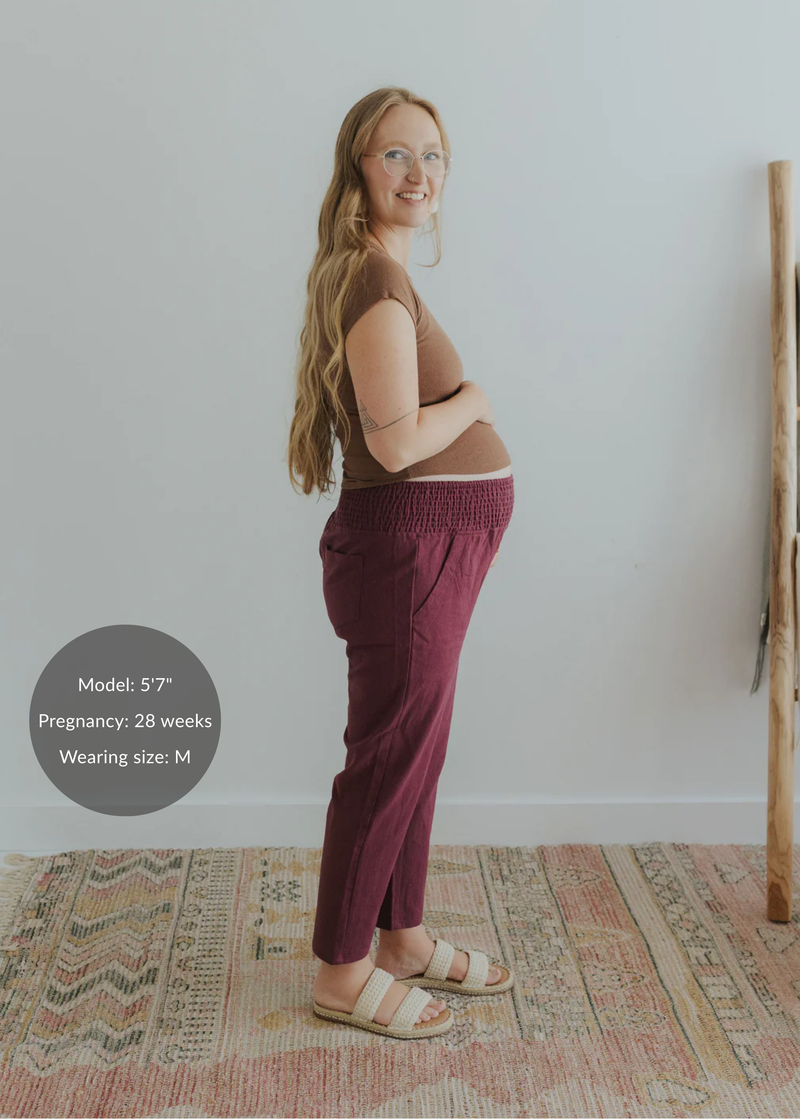 Smiling pregnant woman in a size M, wearing burgundy smocked linen maternity pants with pockets.