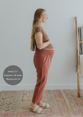 Pregnant woman in rust smocked linen pants with pockets, brown top, and woven sandals.