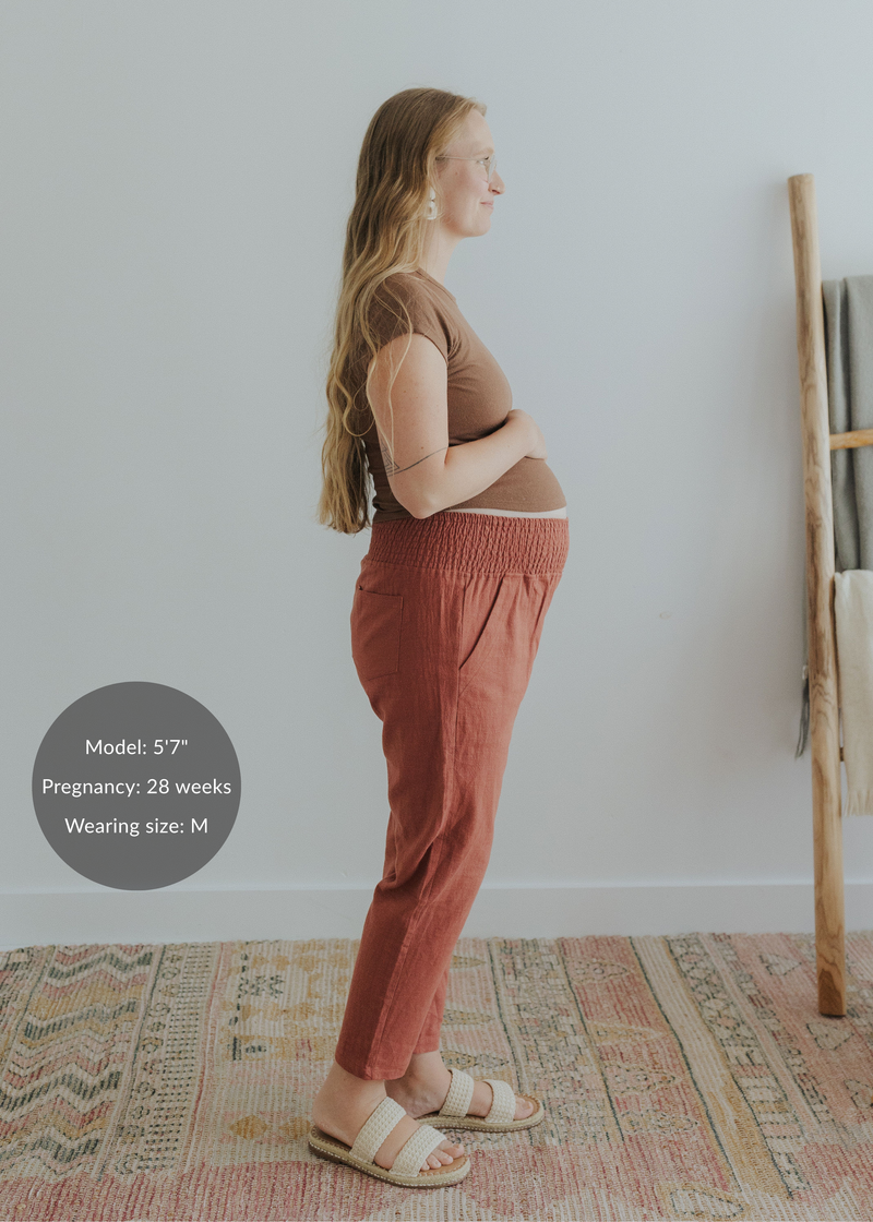 Pregnant woman in rust smocked linen pants with pockets, brown top, and woven sandals.