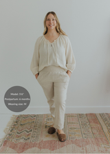 Smiling woman in natural linen smocked maternity pants and matching top, standing on a patterned rug.