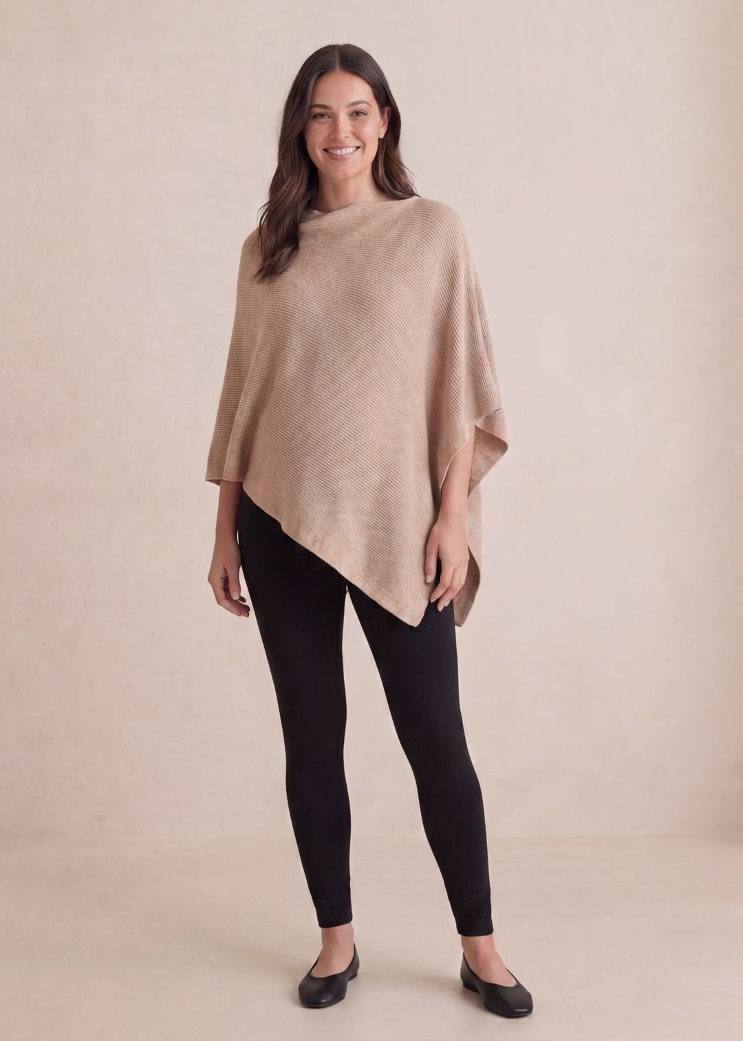 Smiling woman in a beige cashmere/wool asymmetrical knit nursing poncho and black leggings.