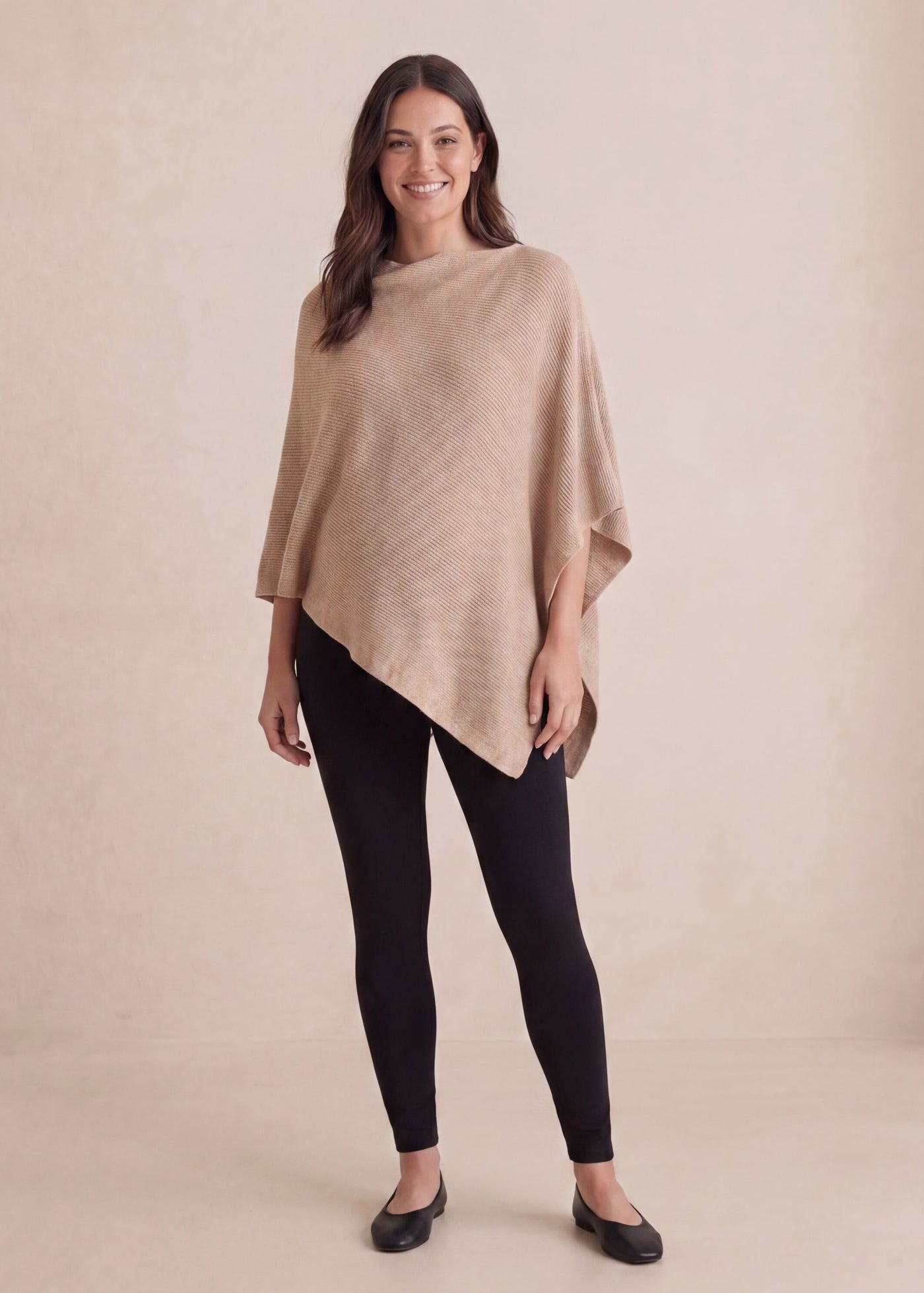 Smiling woman in a beige cashmere/wool asymmetrical knit nursing poncho and black leggings.
