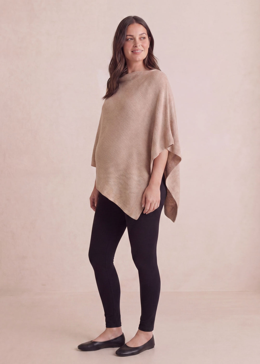 Smiling woman in a beige asymmetrical knit cashmere/wool nursing poncho and black leggings.
