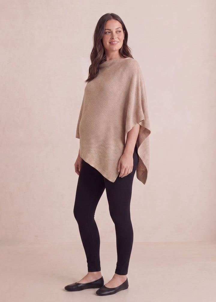 Smiling woman in a beige asymmetrical knit cashmere/wool nursing poncho and black leggings.