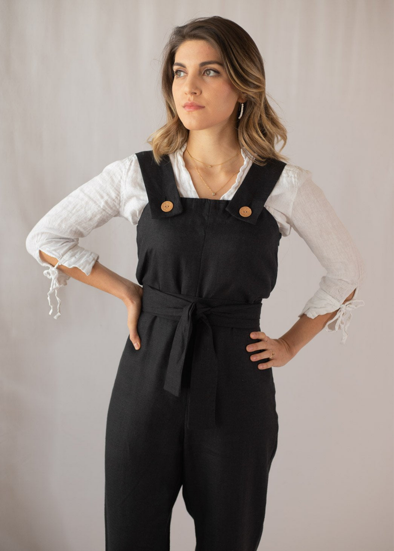 Woman in black linen Beatrice Button Jumpsuit with wood buttons and tie waist over a white long-sleeve shirt.