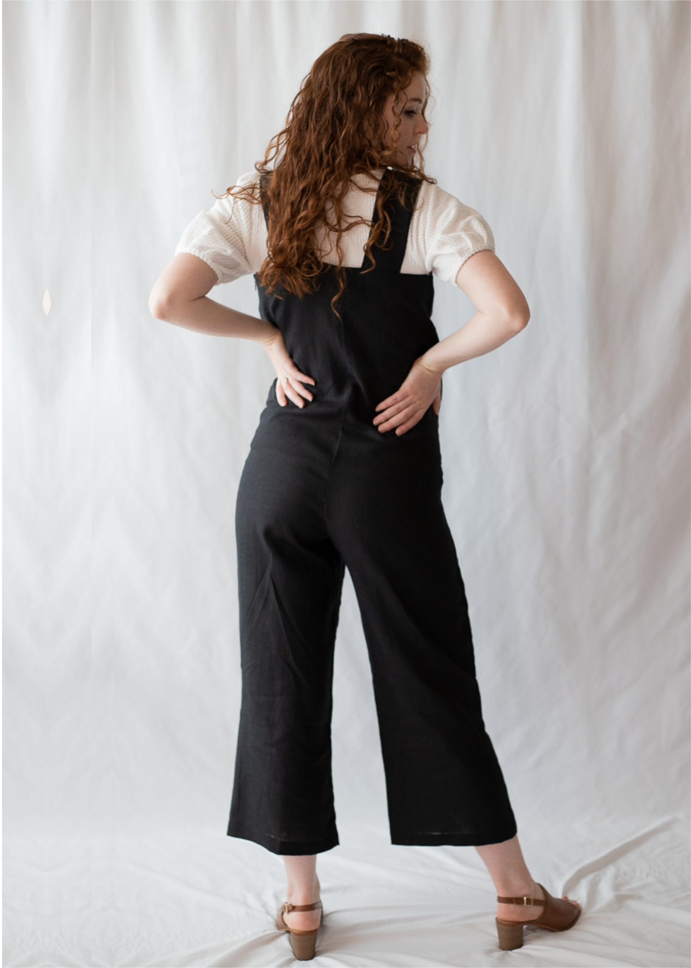 Woman from behind wearing a black linen Beatrice Button Jumpsuit over a white shirt, with brown sandals.