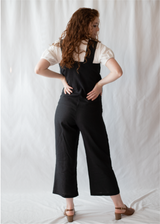 Woman from behind wearing a black linen Beatrice Button Jumpsuit over a white shirt, with brown sandals.