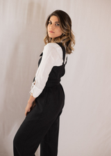 Woman in a black linen Beatrice Button Jumpsuit over a white long-sleeve shirt, looking over her shoulder.