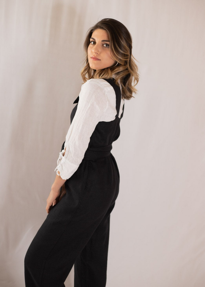 Woman in a black linen Beatrice Button Jumpsuit over a white long-sleeve shirt, looking over her shoulder.