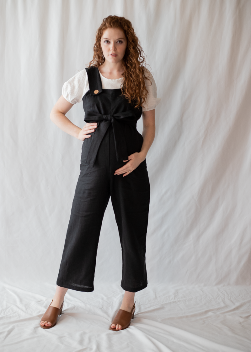 Pregnant woman in a black linen Beatrice Button Jumpsuit with a tie waist and white top.