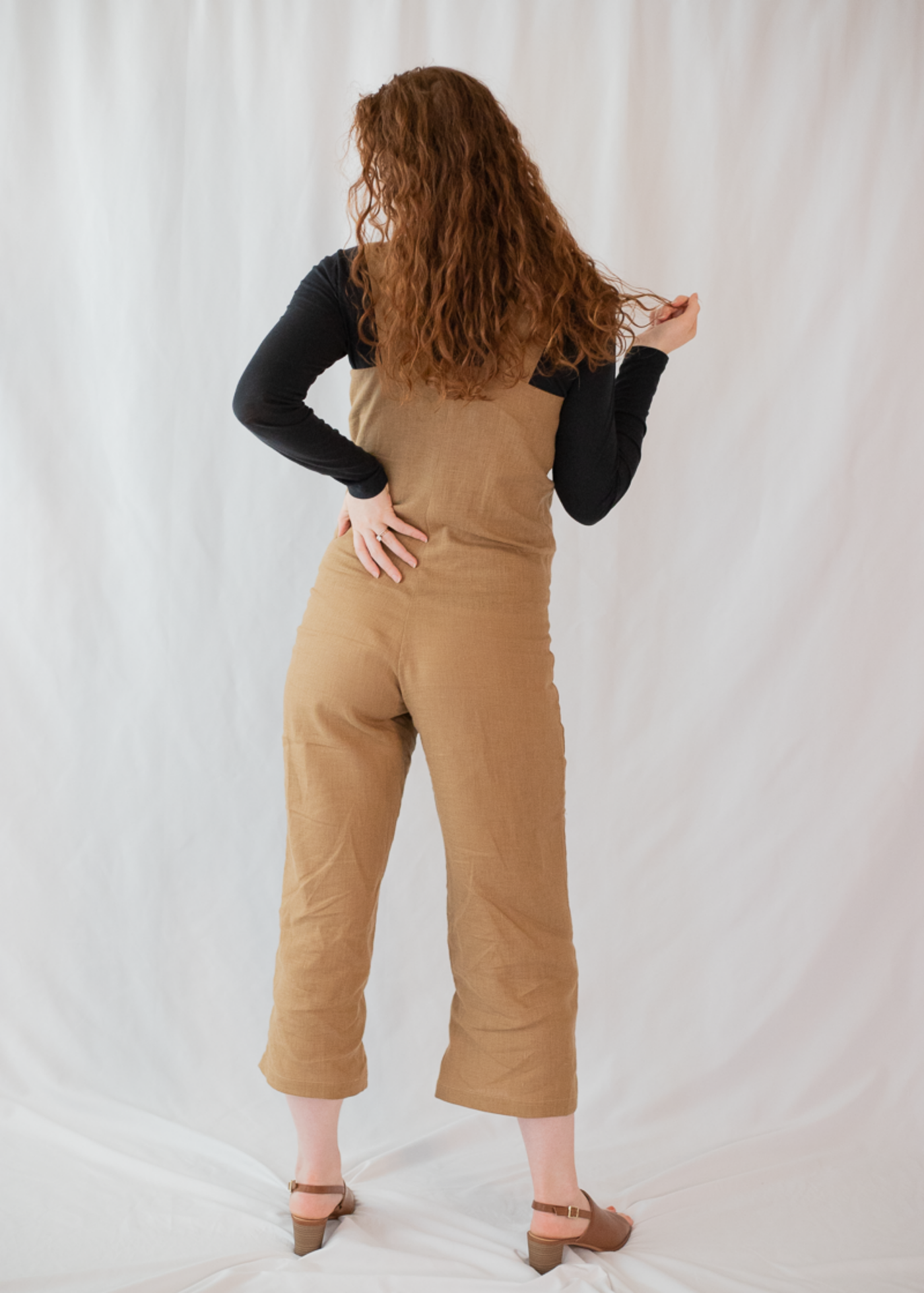 Back view of a woman in a Beatrice Button Jumpsuit, a brown linen maternity jumpsuit over a black long-sleeve shirt.