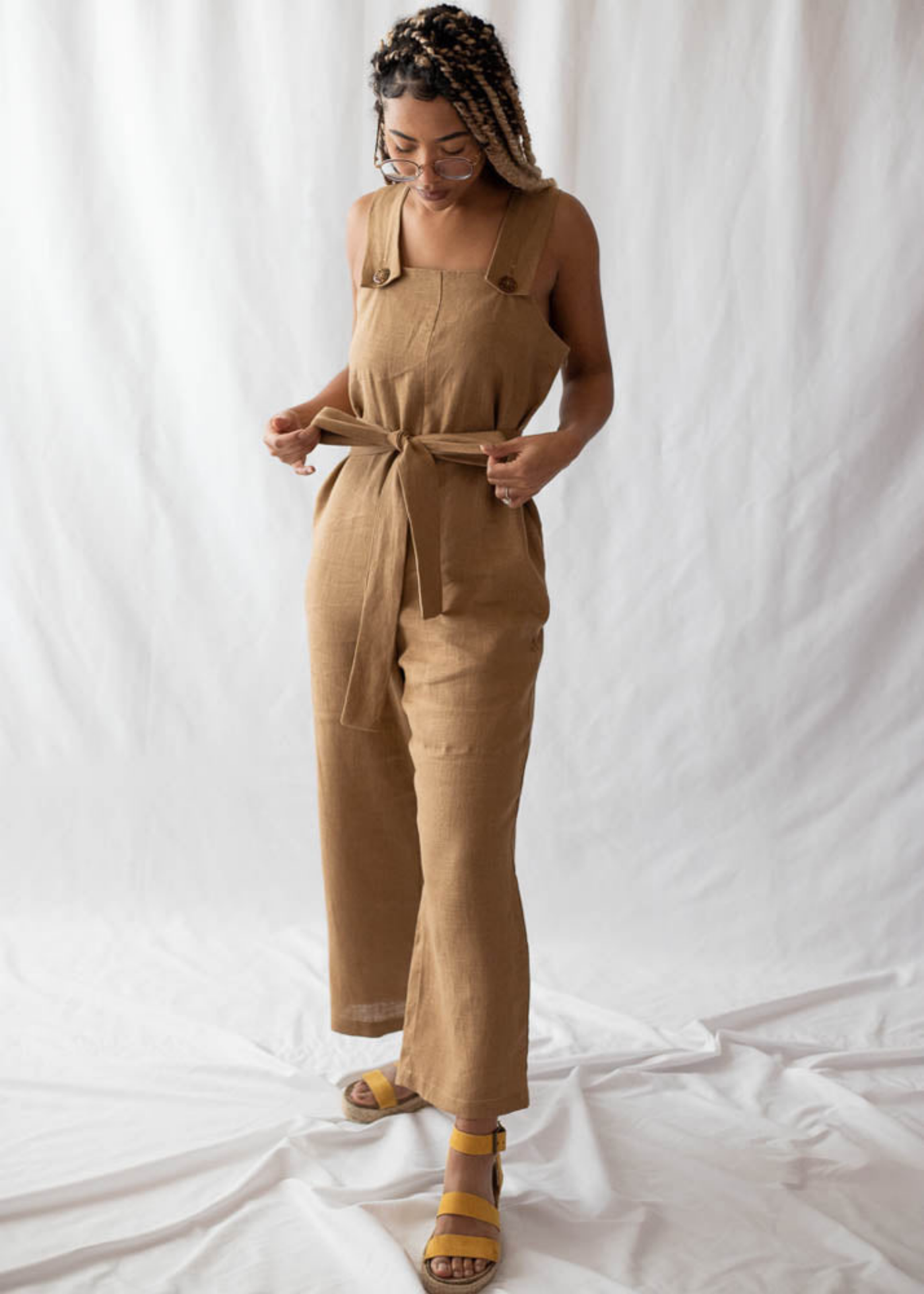 Woman in a Beatrice Button Jumpsuit: a relaxed-fit, tan linen jumpsuit with a tied waist and button straps.