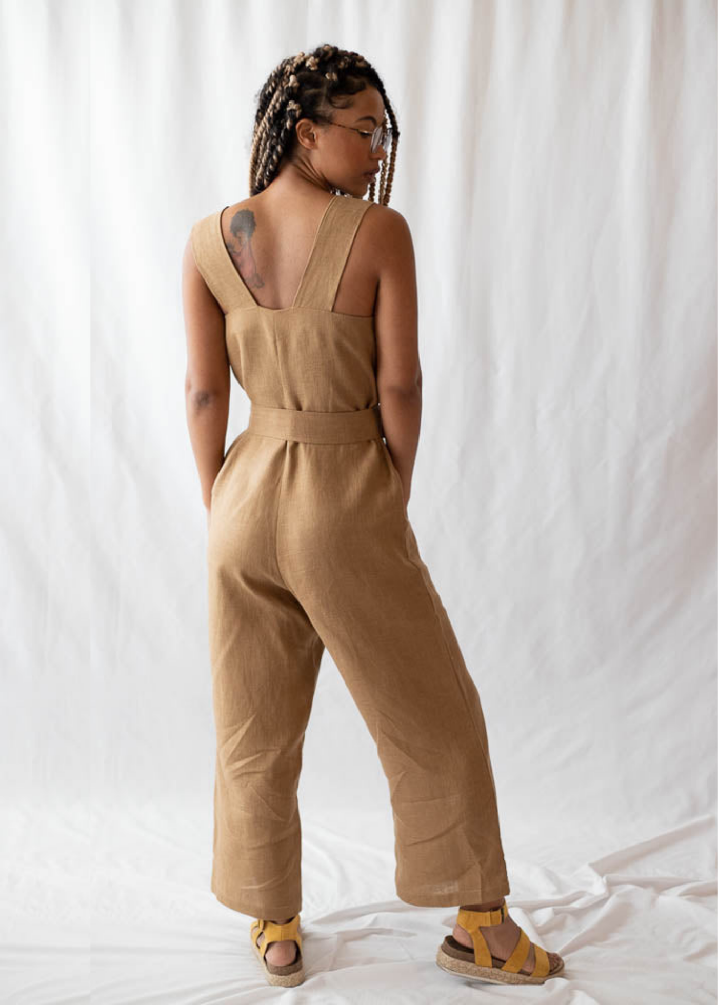 Woman in a back view wearing a tan linen Beatrice Button Jumpsuit with a belted waist and wide legs.