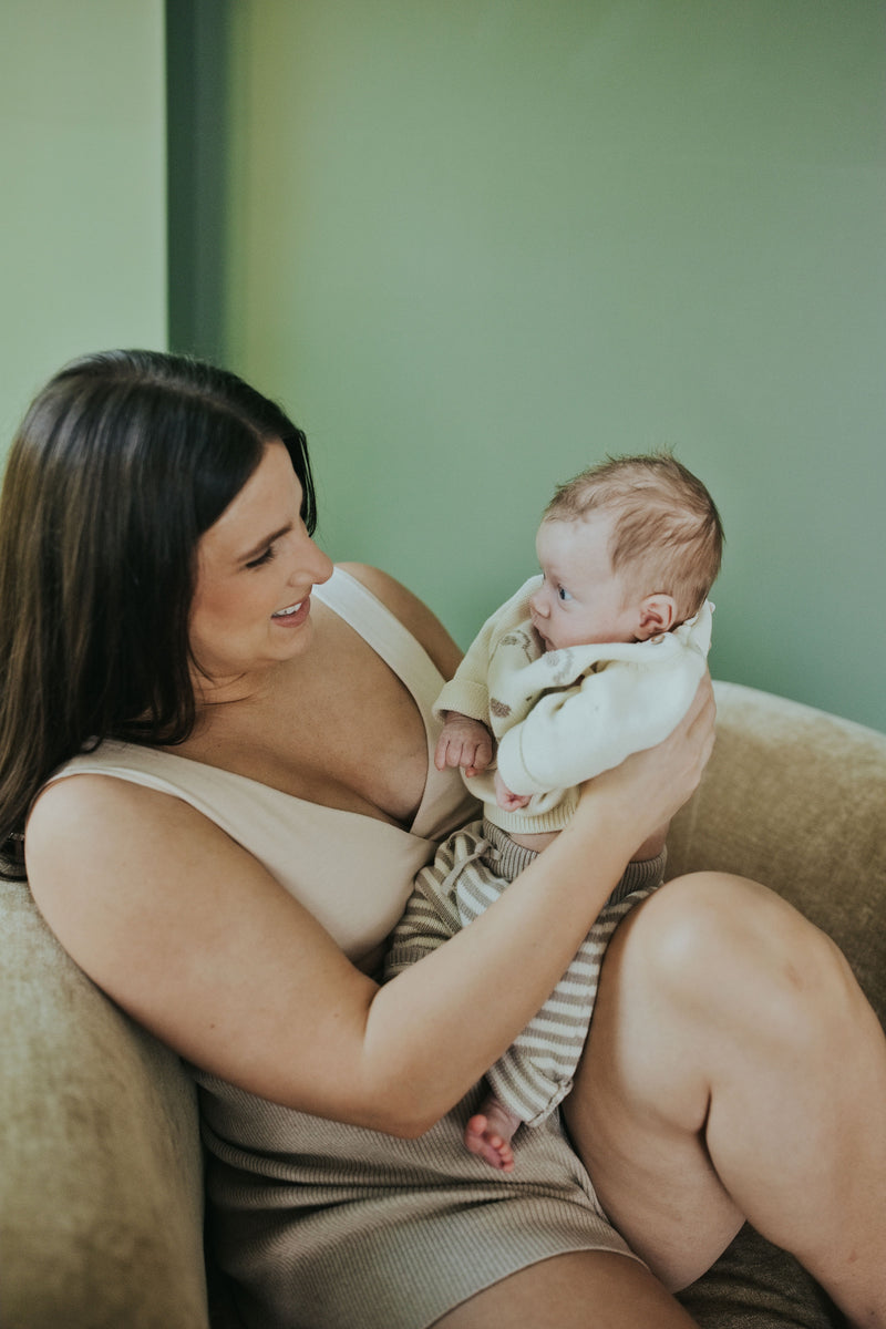 Woman holding her baby in the Sand Crossover Sleep Bra, seated
