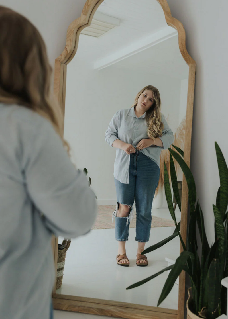 Woman in light blue oversized cotton/linen button-down shirt and ripped jeans, reflected in a wooden mirror.