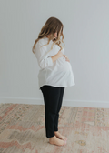 Pregnant woman in a white oversized cotton/linen button-down shirt and black pants, cradling her bump.