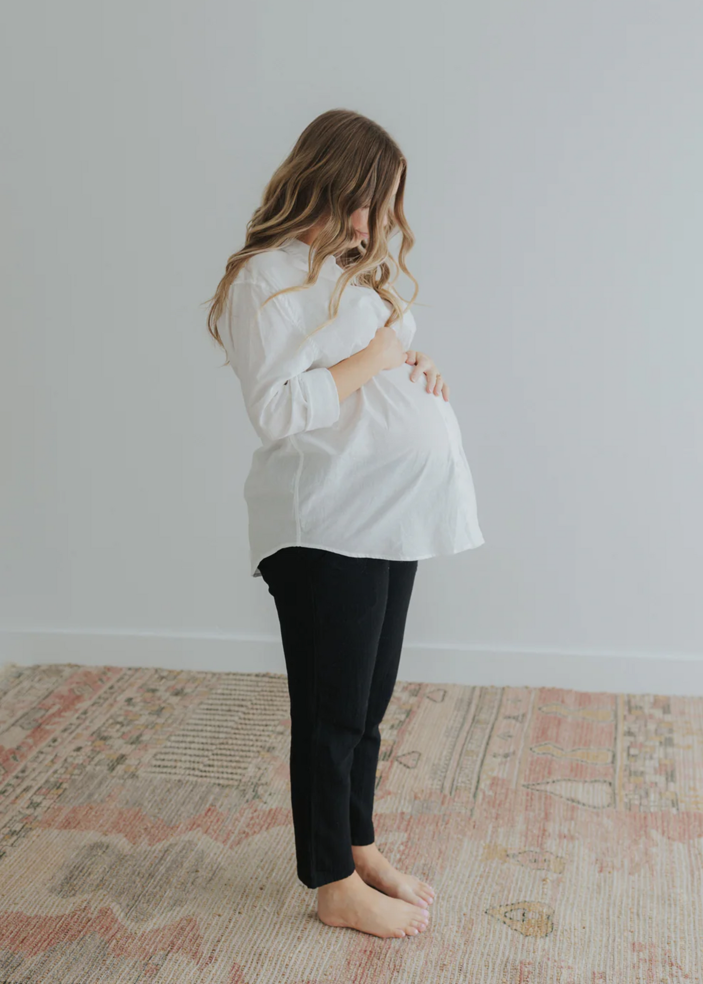 Pregnant woman in a white oversized cotton/linen button-down shirt and black pants, cradling her bump.