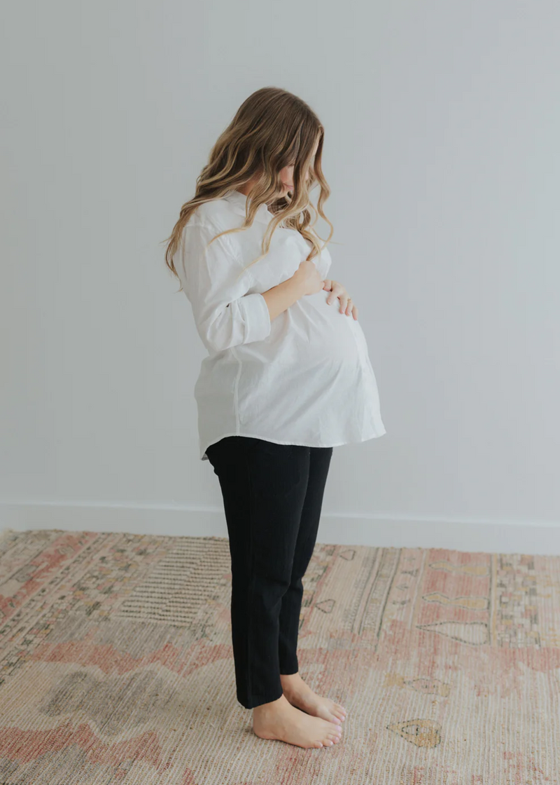 Pregnant woman in a white oversized cotton/linen button-down shirt and black pants, cradling her bump.