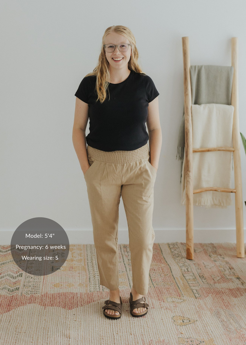 Smiling woman in tan smocked linen maternity pants with pockets and a black tee.