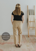 Woman from behind wearing tan smocked linen maternity pants with pockets and a black top.