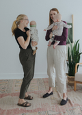 Two women, one in olive green and one in natural linen smocked maternity pants, each holding a baby.