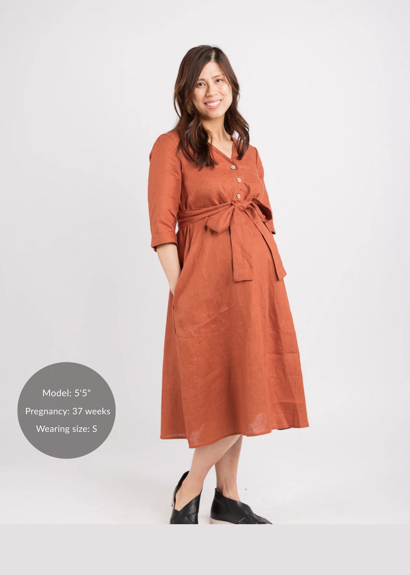 Smiling pregnant woman in a rust linen Faye Dress with front buttons, V-neck, waist tie, and side pockets.