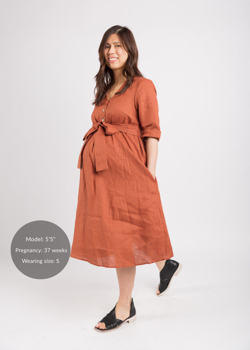 Pregnant model in a rust linen Faye Dress with front buttons, V-neck, waist tie, and side pockets.