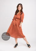 Smiling pregnant model in a rust linen Faye Dress with a V-neck, front buttons, waist tie, and side pockets.