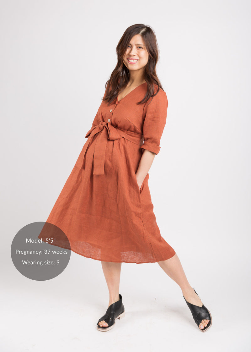 Smiling pregnant model in a rust linen Faye Dress with a V-neck, front buttons, waist tie, and side pockets.
