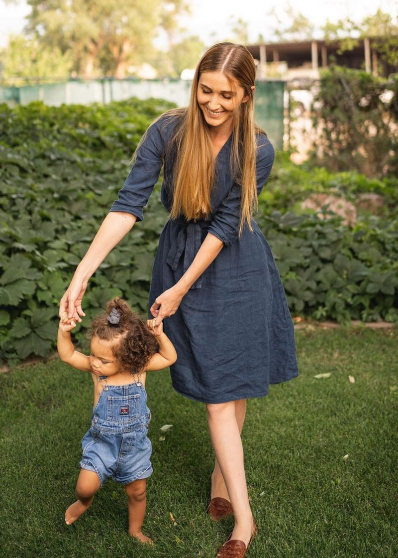 Smiling woman in a navy linen Faye Dress with a tie waist, V-neck, and buttons, holding a toddler's hands.